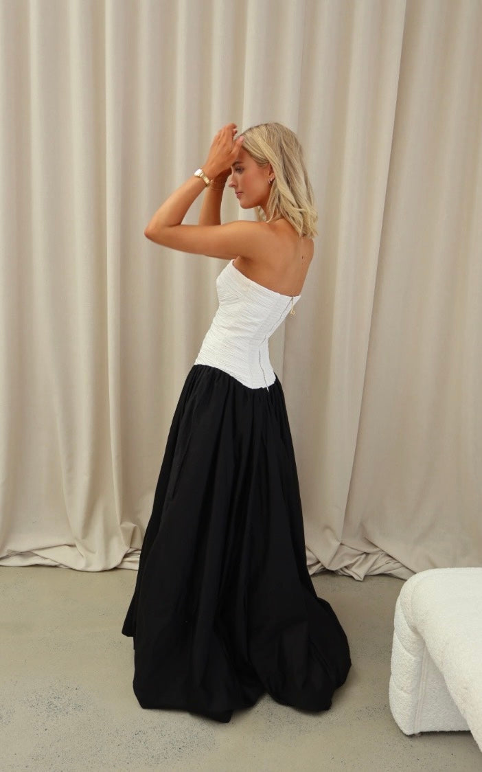 Aje Violette Bubble Hem dress in BLack and White. Model satnds side on with arms raised and hands touching her forehead. The blonde model stands in front of an off white curtain. 