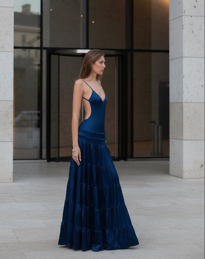 A woman modeling a dark navy blue satin Fenity Raven maxi dress with spaghetti straps, a side cutout, and a tiered skirt, photographed against a modern building entrance. 