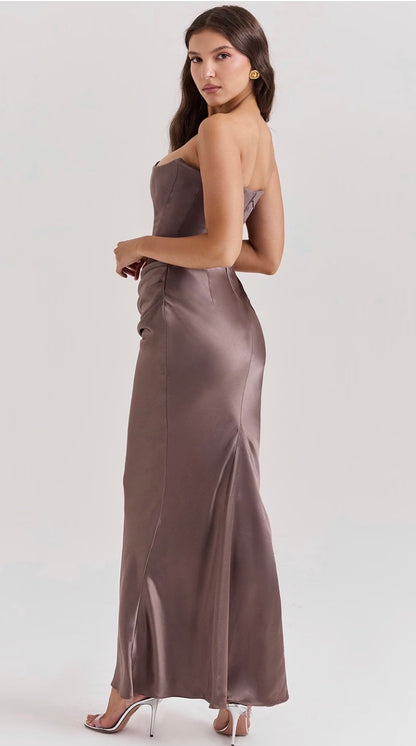 A woman modeling a strapless satin maxi dress in a taupe-brown color, shown from a three-quarter back angle against a light gray studio background.