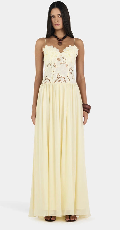 A woman modeling a sleeveless, floor-length cream butter yellow Hansen and Gretel Stephanie gown with a sheer floral lace bodice and a flowing chiffon-like skirt, photographed against a plain light gray studio background.