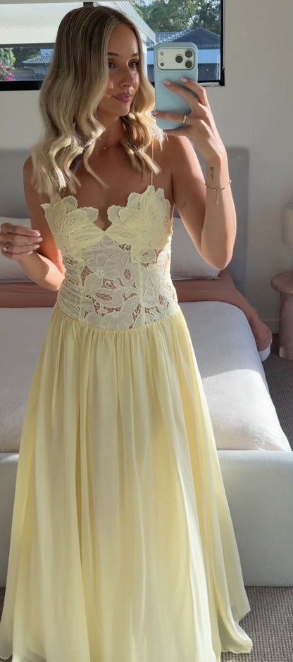 A woman takes a mirror selfie in a bedroom wearing a pale yellow strapless gown with a floral lace bodice and a flowing chiffon skirt.