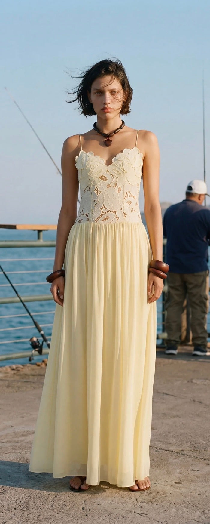 A woman wearing a butter yellow lace and chiffon maxi dress stands on a concrete pier near the water, with a fisherman visible in the background.