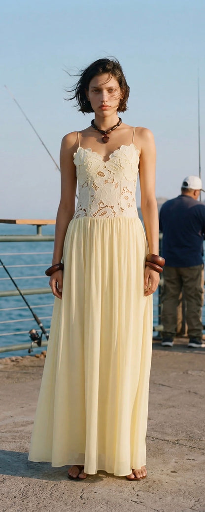 A woman wearing a butter yellow lace and chiffon maxi dress stands on a concrete pier near the water, with a fisherman visible in the background.