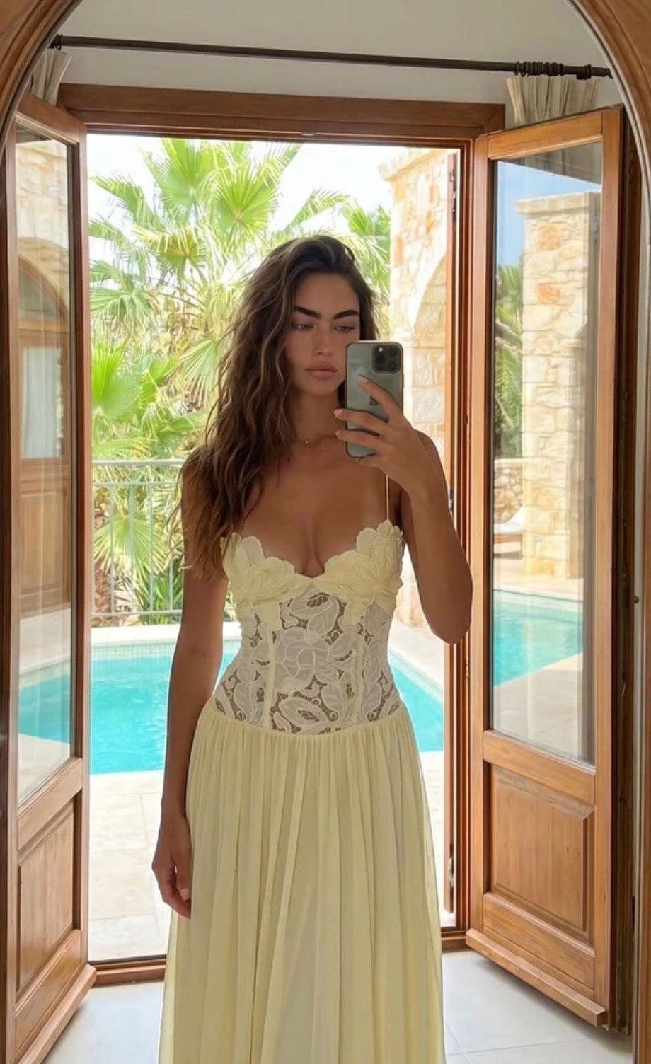 A woman takes a mirror selfie wearing a butter yellow strapless maxi dress with a lace corset-style bodice and a flowing skirt, standing in front of open wooden doors that lead to a poolside patio.