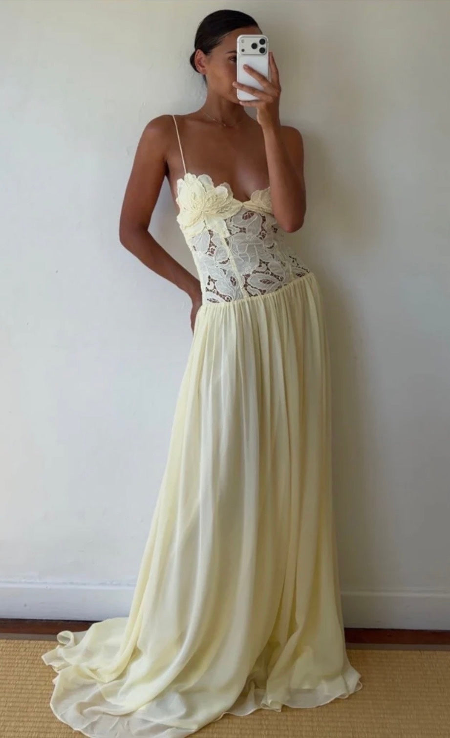 A model takes a mirror selfie wearing a long butter yellow Hansen and Gretel Stephanie gown with a lace corset bodice and a flowing chiffon skirt, standing against a plain off-white wall.