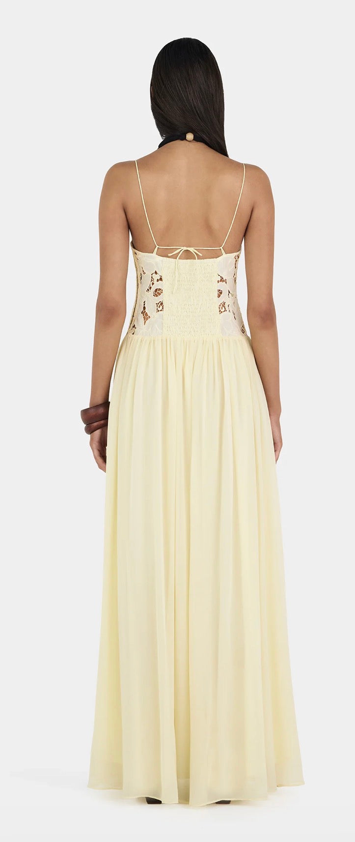 A woman viewed from the back wearing a long yellow Hansen and Gretel Stephanie maxi dress with thin straps, a lace-up open back, and embroidered detailing on the upper back, against a plain light gray studio background. 