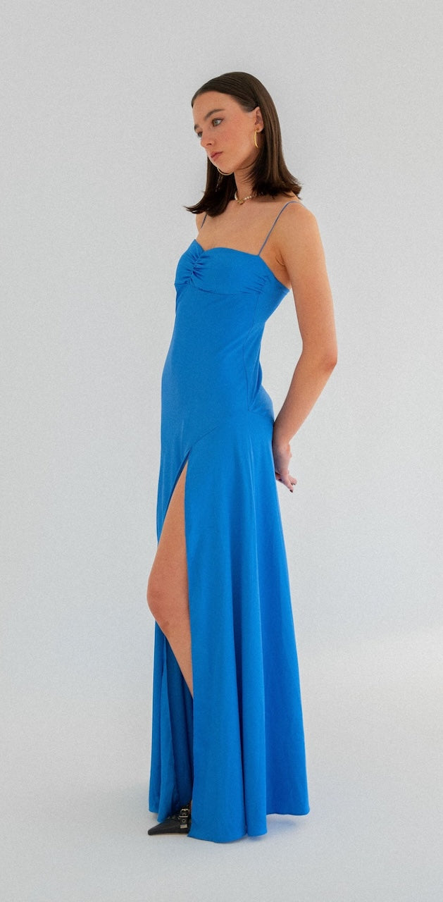 Hntr Gaia Gown Azul side view. Photo shows model wearing blue dress with a leg split looking away with a white background. 