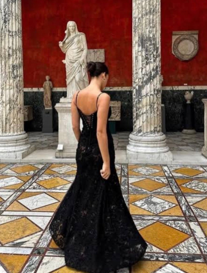 Woman in a black House of CB Seren dress standing in an elegant interior setting with classical columns and statues.