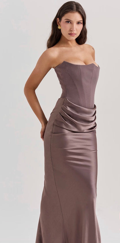 A woman modeling a House of CB strapless Persephone satin evening gown in a muted taupe-brown color against a light gray studio background.