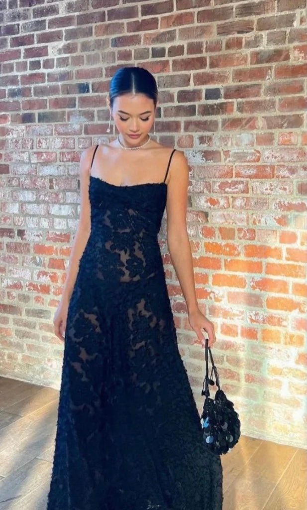 Woman in a black lace House of CB Seren dress holding a black handbag against a brick wall.